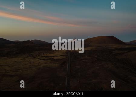 Atemberaubender Blick auf die Asphaltstraße durch wildes, raues Gelände mit felsigen Bergen vor dem Sonnenuntergang am Himmel auf Lanzarote Stockfoto