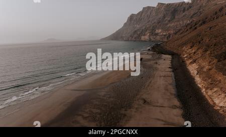 Malerischer Blick auf die Wellen am Sandstrand vor felsigen Felsformationen unter dem bewölkten Sonnenuntergang auf Lanzarote Stockfoto