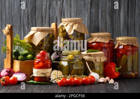 Eingelegte Gurken in einem Glas, eine Holzkiste mit Konservierung auf dunklem Holzhintergrund. Paprika, Zwiebeln und frische Cornichons zur Konservierung. Stockfoto