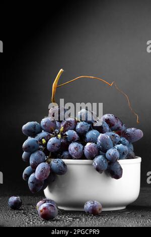 Ein Haufen blauer Trauben auf einem weißen Teller auf dunklem Hintergrund, mit Trauben auf dem Tisch verstreut. Ein Haufen Trauben mit Wassertropfen vertikal Stockfoto