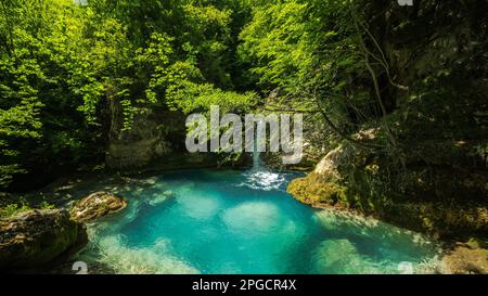 Kleiner Wasserfall, der in einen blauen See fließt, umgeben von Felsen und grünen Bäumen, die an sonnigen Tagen im Wald wachsen Stockfoto
