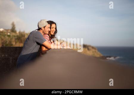 Seitenansicht eines jungen Paares in legerer Kleidung und Kappe, das sich auf einen Steinbock lehnt und auf das blaue Meer blickt, während Sie die Küste der Azoren, Portugal, genießen Stockfoto