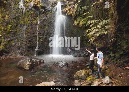 Seitenansicht eines jungen Paares, das bei Tageslicht in der Azoren-Region in der Nähe des Wasserfalls im grünen Wald Händchen hält Stockfoto