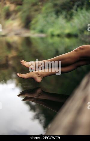 Barfuß-Beine einer anonymen Reisenden, die am Rande eines ruhigen Teiches in der Natur sitzt Stockfoto