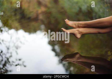 Barfuß-Beine einer anonymen Reisenden, die am Rande eines ruhigen Teiches in der Natur sitzt Stockfoto