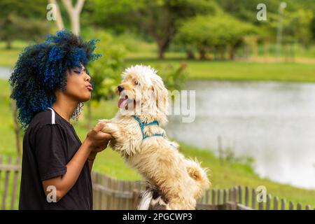 Goiania, Goias, Brasilien – 20. März 2023: Eine junge schwarze Frau mit gefärbtem blauem Haar, mit ihrem Hund auf dem Schoß, in einem bewaldeten Park. Stockfoto