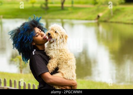 Goiania, Goias, Brasilien – 20. März 2023: Eine junge schwarze Frau mit gefärbtem blauem Haar, mit ihrem Hund auf dem Schoß, in einem bewaldeten Park. Stockfoto