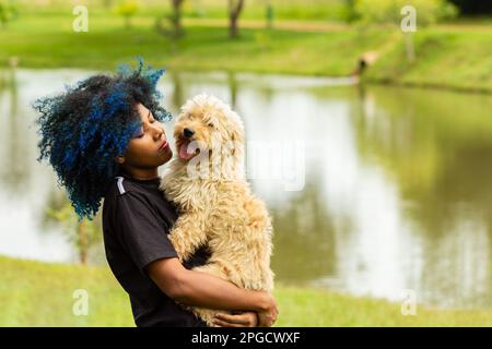 Goiania, Goias, Brasilien – 20. März 2023: Eine junge schwarze Frau mit gefärbtem blauem Haar, mit ihrem Hund auf dem Schoß, in einem bewaldeten Park. Stockfoto