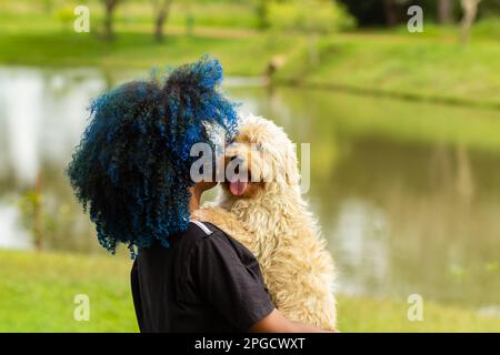 Goiania, Goias, Brasilien – 20. März 2023: Eine junge schwarze Frau mit gefärbtem blauem Haar, mit ihrem Hund auf dem Schoß, in einem bewaldeten Park. Stockfoto