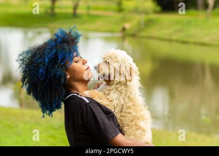 Goiania, Goias, Brasilien – 20. März 2023: Eine junge schwarze Frau mit gefärbtem blauem Haar, mit ihrem Hund auf dem Schoß, in einem bewaldeten Park. Stockfoto