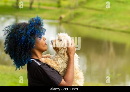 Goiania, Goias, Brasilien – 20. März 2023: Eine junge schwarze Frau mit gefärbtem blauem Haar, mit ihrem Hund auf dem Schoß, in einem bewaldeten Park. Stockfoto