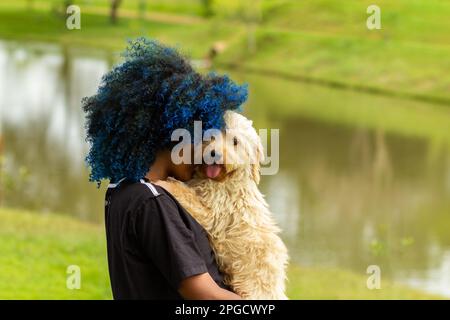 Goiania, Goias, Brasilien – 20. März 2023: Eine junge schwarze Frau mit gefärbtem blauem Haar, mit ihrem Hund auf dem Schoß, in einem bewaldeten Park. Stockfoto