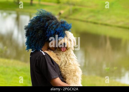 Goiania, Goias, Brasilien – 20. März 2023: Eine junge schwarze Frau mit gefärbtem blauem Haar, mit ihrem Hund auf dem Schoß, in einem bewaldeten Park. Stockfoto