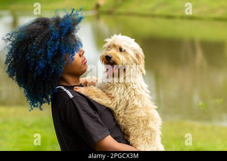 Goiania, Goias, Brasilien – 20. März 2023: Eine junge schwarze Frau mit gefärbtem blauem Haar, mit ihrem Hund auf dem Schoß, in einem bewaldeten Park. Stockfoto