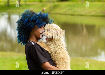 Goiania, Goias, Brasilien – 20. März 2023: Eine junge schwarze Frau mit gefärbtem blauem Haar, mit ihrem Hund auf dem Schoß, in einem bewaldeten Park. Stockfoto