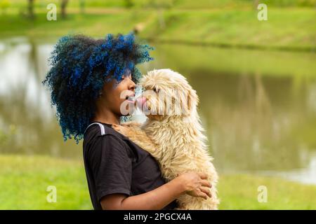 Goiania, Goias, Brasilien – 20. März 2023: Eine junge schwarze Frau mit gefärbtem blauem Haar, mit ihrem Hund auf dem Schoß, in einem bewaldeten Park. Stockfoto