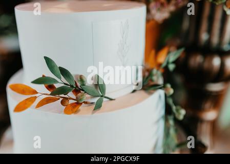 Ein appetitlicher weißer Kuchen mit grünem Laub und bunten Blumen auf dem Oberteil, bereit zum Servieren Stockfoto