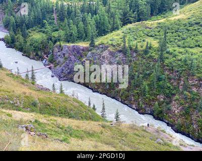 Ein Tourist von einer Klippe aus blickt auf eine kleine hölzerne Brücke aus Brettern über den Berg Fluss Chuya mit Fluss und schlammigem Wasser zwischen den Felsen mit sp Stockfoto