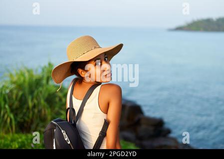 Eine multiethnische Frau mit Strohhut genießt einen tropischen Urlaub auf einer Bank an der Klippe mit Blick auf das Meer. Schwarze Frau mit Rucksack für Besichtigungstouren auf landschaftlich reizvoller Landschaft Stockfoto