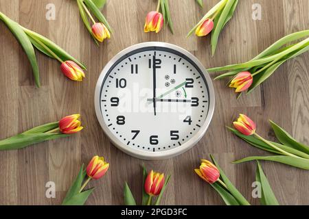 Eine Uhr zeigt drei Stunden an. Tulpen liegen herum. Ein Symbol für die Zeitänderung. Sommerzeit. Bewegen Sie die Hände nach vorne. Der grüne Pfeil ist drin Stockfoto