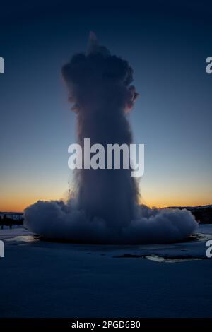 Islands großer Geysir Strokkur im vollen Ausbruch mit Nebel und Rauch, Hintergrundbeleuchtung und dem letzten Licht des orangefarbenen Sonnenuntergangs oder der nahe Dämmerung dahinter Stockfoto