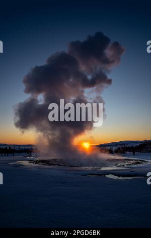 Islands großer Geysir Strokkur in voller Eruption mit Nebel und Rauch von hinten beleuchtet und die orangefarbene Abendsonne oder die nahe Dämmerung berührt den Horizont dahinter Stockfoto