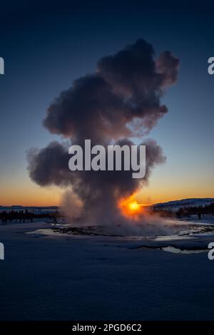 Islands großer Geysir Strokkur in voller Eruption mit Nebel und Rauch von hinten beleuchtet und die orangefarbene Abendsonne oder die nahe Dämmerung berührt den Horizont dahinter Stockfoto