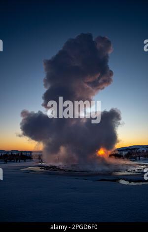 Islands großer Geysir Strokkur im vollen Ausbruch mit Nebel und Rauch, Hintergrundbeleuchtung und dem letzten Licht des orangefarbenen Sonnenuntergangs oder der nahe Dämmerung dahinter Stockfoto