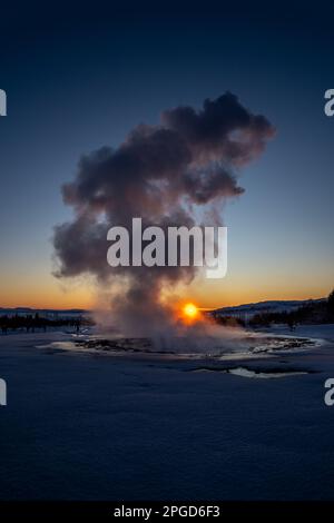 Islands großer Geysir Strokkur in voller Eruption mit Nebel und Rauch von hinten beleuchtet und die orangefarbene Abendsonne oder die nahe Dämmerung berührt den Horizont dahinter Stockfoto