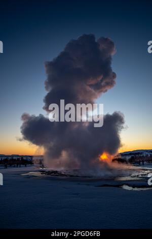 Islands großer Geysir Strokkur in voller Eruption mit Nebel und Rauch von hinten beleuchtet und die orangefarbene Abendsonne oder die nahe Dämmerung berührt den Horizont dahinter Stockfoto