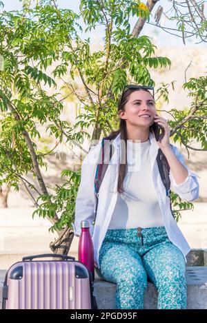 Weibliche Reisende, die mit Rucksack und Koffer im Freien am Telefon sitzt. Stockfoto