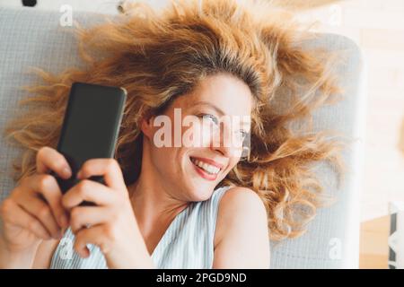 Nahaufnahme des Porträts einer Frau mit lockigem Haar, die wegschaut und auf der Couch liegt, während sie ihr Telefon hochhält und Selfies macht Stockfoto