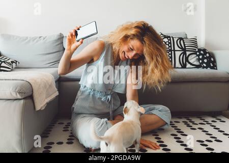 Fröhliche weiße Frau mit lockigen Haaren, die mit der kleinen chihuahua auf dem Teppich in ihrer Wohnung spielt, während sie Selfies mit ihrem Handy macht Stockfoto