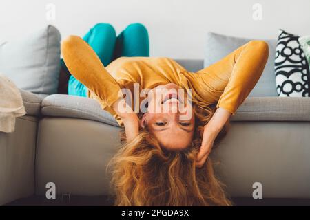 Lächelnde, fröhliche Frau, die auf der Couch lag und ihren Kopf abhing, lächelte die Kamera an und trug einen roten Lippenstift Stockfoto