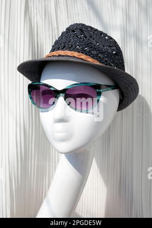 Weibliche Schaufensterpuppe mit blauer Sonnenbrille und Weidenhut im Verkauf in einem Boutique-Fenster. Damenmode-Accessoires für den Sommer zum Schutz vor Stockfoto