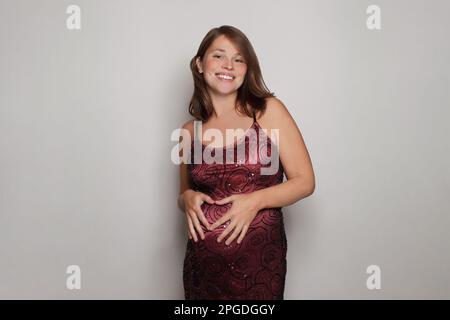 Eine attraktive, lächelnde schwangere Frau, die ein Herz aus der Hand auf dem Bauch macht, während sie vor dem grauen Hintergrund des Studiowandschilds steht Stockfoto
