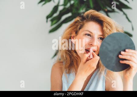Warte auf das Porträt einer Frau, die ihren Lippenstift macht und den kleinen runden Spiegel ansieht, den sie hochhält Stockfoto