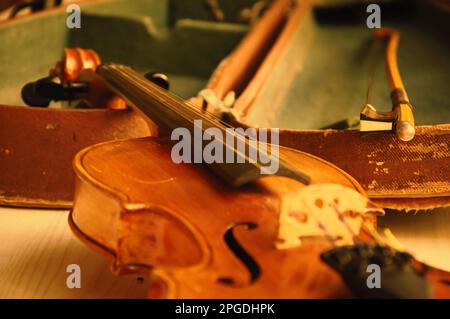 Alte Geige und Schleife, die über einem offenen, verschlissenen Koffer lagen. Selektiver Fokus auf der Bogenspitze. Gealtertes Farbfoto. Stockfoto