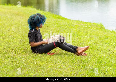 Goiania, Goias, Brasilien – 20. März 2023: Eine junge schwarze Frau mit gelocktem, blau gefärbtem Haar, die auf dem Gras in einem Park sitzt. Stockfoto