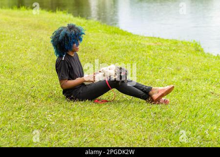 Goiania, Goias, Brasilien – 20. März 2023: Eine junge schwarze Frau mit gelocktem, blau gefärbtem Haar, die auf dem Gras in einem Park sitzt. Stockfoto