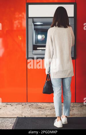 Vertikales Foto, Rückansicht einer Frau, die vor einem Geldautomaten an einer roten Wand steht und Bargeld abhebt Stockfoto