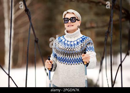 Glückliche Frau, nordic, die mit Stöcken im Naturwald spaziert. Sportlicher, aktiver Lifestyle. Weibliches Modell im Außenporträt mit kurzen blonden Haaren in Sonnenbrille und Stockfoto