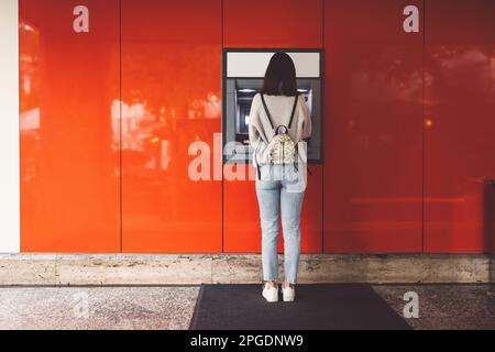 Rückansicht einer jungen Frau, die vor einem Geldautomaten an einer roten Wand steht Stockfoto