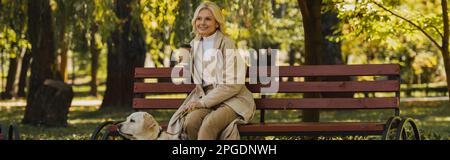 Fröhliche blonde Frau, die Kaffee zum Mitnehmen hält, während sie Zeit mit labrador im Park verbringt, Banner, Stock Image Stockfoto