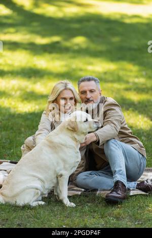 Glückliches Paar mittleren Alters, das labrador-Hund kuschelte, während es beim Picknick im Park auf der Decke saß, Stockbild Stockfoto
