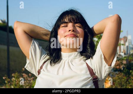 Machen Sie eine Pause von einer langen Klasse an der Universität, junge Frau im Freien, sitzt mit Armen hinter dem Kopf und Augen geschlossen, entspannt und atmet. Stockfoto