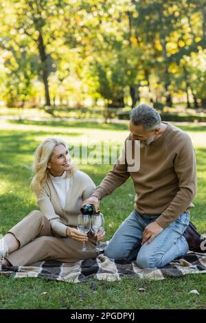 Ein Mann mittleren Alters, der Wein in das Glas schüttet, neben einer fröhlichen Frau während des Picknickens im Park, Bild der Börse Stockfoto