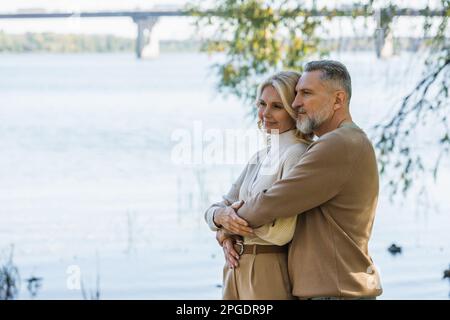 Ein fröhlicher Mann mittleren Alters mit grauem Bart umarmt eine glückliche blonde Frau in der Nähe des Flusses im Park, Stockbild Stockfoto