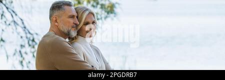 Ein fröhlicher Mann mittleren Alters mit grauem Bart umarmt eine glückliche blonde Frau in der Nähe des Flusses, Banner, Stock Image Stockfoto