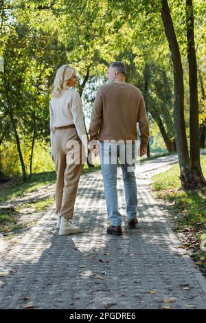 Ein stilvolles Paar mittleren Alters in voller Länge, das Händchen hält, während es im grünen Park spaziert, Stockbild Stockfoto